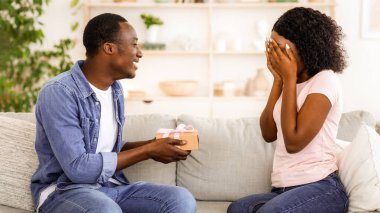 Afrikalı bir adam kız arkadaşına hediye paketi veriyor. Kadın avuçlarıyla gözlerini kapatıyor. Boş bir alan.