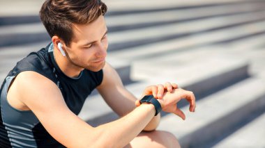 Modern koşu aletleri. Kablosuz kulaklıklı yorgun sporcu fitness izleyicisine bakar ve basamaklara oturur, kapalı, boş alan, güneş parlaması