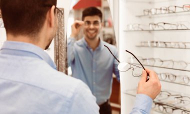 Bir adam bir optik mağazada dikiliyor, bir çift gözlük denerken yansımasını inceliyor. Yeni görünüşünü düşünürken gülümsemesi memnuniyeti yansıtıyor..