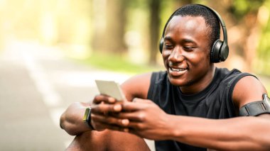 Mola sırasında akıllı telefon kullanan, parkta antrenman yapan, ücretsiz alan kullanan, kablosuz kulaklıklı gülümseyen Afrikalı sporcunun yakın çekimi.