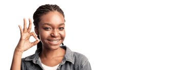 Genç bir bayan sağ eliyle iyi bir el hareketi yaparken geniş bir şekilde gülümsüyor. Kendinden emin ve mutlu görünüyor. Aydınlatılmış bir yerde günlük giysiler giyiyor..