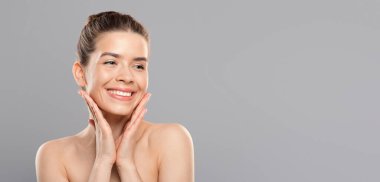 Gri stüdyo arka planında gülümseyen şehvetli genç bir kadın yüzüne dokunuyor ve fotokopi odasına bakıyor. Kadın güzellik anlayışı, panorama. Gri üzerine çıplak, mutlu bayan reklam için yer var.