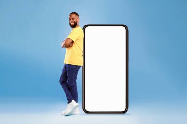 Neşeli bir yüzü olan bir adam büyük, boş bir akıllı telefonun yanında güvenle durur. Parlak sarı bir gömlek ve rahat bir pantolon giyiyor. Canlı bir stüdyo ortamında modern bir yaşam tarzı sergiliyor..