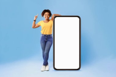 Genç bir Afro-Amerikalı kadın, boş bir akıllı telefonun yanında neşeli bir ifadeyle, kendi tarzını izole bir ortamda sergiliyor. Bu modern bir estetik yaşam tarzını yakalar..