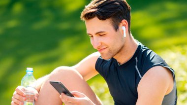 Spor için modern teknolojiler. Kablosuz kulaklıklı gülen adam akıllı telefona bakar ve yeşil çimen arka planında bir şişe su tutar, panorama, serbest alan