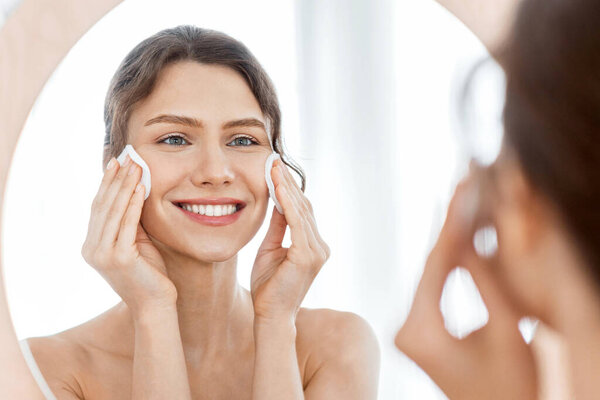 Skincare. Young cheerful woman looking at mirror and cleaning her face with cotton pads at home