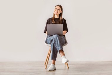 Genç esmer bir kadın dizüstü bilgisayarıyla meşgul bir şekilde koltukta oturuyor. Günlük kıyafetler giyiyor ve rahat görünüyor. Minimalist bir iç mekan ortamında bin yıllık bir yaşam tarzını yansıtıyor..