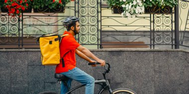 Genç adam şehirde paket teslimatı yapan bir bisiklet sürüyor. Sokak, profil, kopyalama alanında sarı sırt çantalı kurye tarafından çevrimiçi sipariş