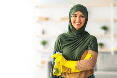 Genç bir Orta Doğulu kadın yeşil bir başörtüsü ve parlak sarı eldivenler giyerek evini neşeyle temizliyor. Elinde bir sprey şişesi tutuyor ve düzenli bir çevreyi korumayı seviyor.