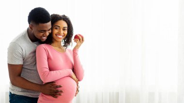 Kocası ona arkadan sarılırken, mutlu hamile Afrikalı kadın elma yiyor.