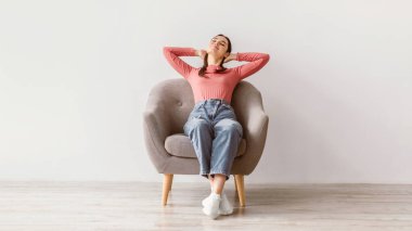 Genç bir esmer kadın rahat bir koltukta otururken bir anlık rahatlığın tadını çıkarıyor. Kollarını başının arkasına uzatıyor şık, minimalist, kapalı bir ortamda, huzurlu bir yaşam tarzını kucaklıyor..