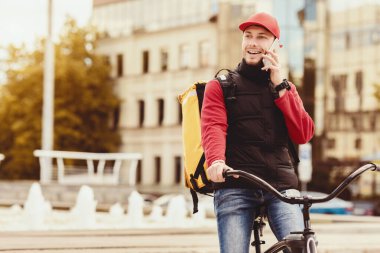 Bisikletin üzerinde oturan, cep telefonuyla konuşan kurye, şehir merkezindeki açık havada yemek dağıtıyor. Boşluğu Kopyala
