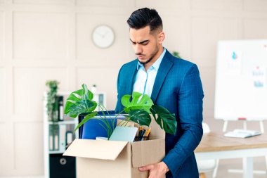 Genç bir Arap girişimci, elinde bitkiler, dosyalar ve ofis malzemeleriyle dolu bir kutu ile modern bir ofiste duruyor ve ayrılmaya hazırlanırken ciddi bir tavır sergiliyor..