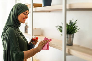 Genç Müslüman bir kadın şık evindeki ahşap rafları temizlemekle meşgul. Başörtüsü takıyor ve tozu silerken gülümsüyor. Yaşam alanının düzenliliğinin tadını çıkarıyor..