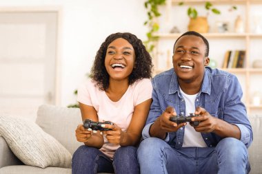 Mutlu Afro-Amerikan çifti evde video oyunu oynuyor, fotokopi uzayı. Neşeli genç siyahi erkek ve kadın oyuncular oturma odasında kanepede oturuyor ve ellerinde joysticklerle rekabet halindeler.
