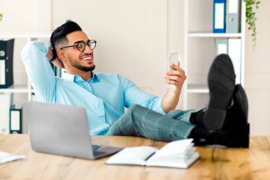 Genç bir Arap işadamı ofisinde dinleniyor, akıllı telefonuna gülümsüyor ve ayakları masanın üzerinde uzanıyor. Yakınlarda bir dizüstü bilgisayar ve not defteri var. Şirket yaşam tarzını yansıtıyor..