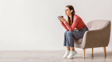 Esmer kadın şık bir koltukta rahatlıyor, içkisini yudumluyor. Düşünceli ve sakin görünüyor, modern bir yaşam tarzını parlak, minimalist bir ortamda somutlaştırıyor..