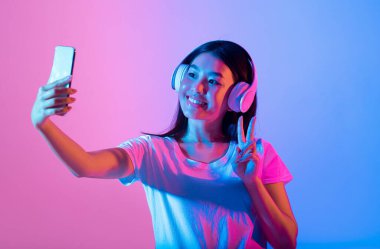 Asyalı kadın akıllı telefonuyla kulaklık takarak selfie çekerken parlak bir şekilde gülümsüyor. Canlı pembe ve mavi ışıklar eğlenceli bir atmosfer yaratır. Poz verirken barış işareti gösteriyor..