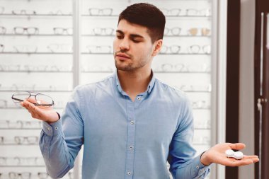 Genç bir adam bir optik mağazada dikiliyor, düşünceli bir şekilde bir elinde gözlük, diğer elinde kontakt lensler, görüş düzeltme seçeneklerini değerlendiriyor..