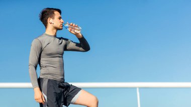 Sokakta yaz antrenmanı. Spor giyim bölümündeki adam mavi gökyüzü arka planındaki stadyumda su içiyor.