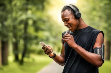 Neşeli Afrikalı sporcu protein yiyor ve dinlenirken akıllı telefon kullanıyor, parkta egzersiz yapıyor, boş alan var.