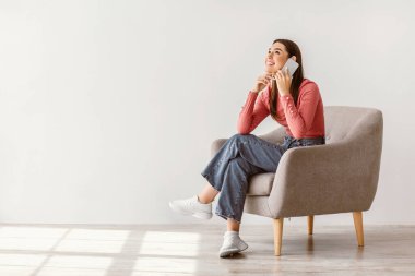 Genç esmer bir kadın şık bir koltukta rahat bir şekilde oturur ve canlı bir telefon görüşmesi yapar. Düşünceli bir şekilde gülümsüyor ve çağdaş yaşam alanına sıcaklık katıyor..
