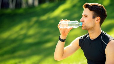 Adam su içiyor. Akıllı saati olan sporcu yeşil park arka planında şişe tutuyor
