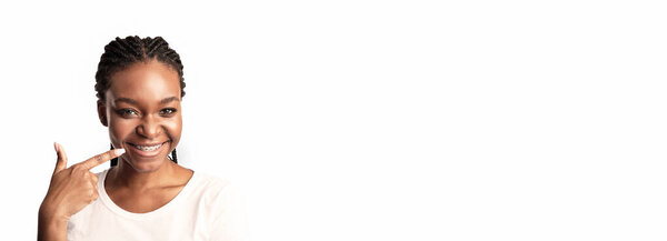 A young woman with braided hair smiles broadly, pointing to her teeth with a playful expression in a bright and minimalistic environment.