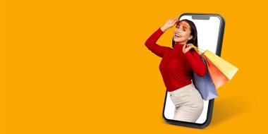 Neşeli bir müşteri çevrimiçi bir mağaza gösteren bir akıllı telefonla etkileşime girer. Elinde alışveriş torbaları var. Evinden alışveriş yapmanın verdiği heyecanı gösteriyor..