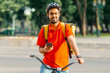 Bisikletli teslimatçı. Sarı sırt çantalı mutlu kurye, akıllı saat ve akıllı telefon, şehirde bisiklete biner, boşluğu kopyalar.