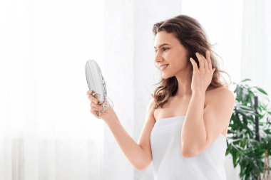 Sabah duşundan sonra havluya sarılı, evde yatakta oturan, aynaya bakan, fotokopi çeken genç kadın. Çekici, gülümseyen bir kadının güzel saçlarına dokunuşunun yan görüntüsü.