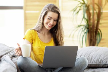 Dizüstü bilgisayarı ve kahvesi olan güzel bir kız yatakta oturuyor ve evden internet üzerinden çalışıyor.