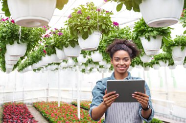 Bahçıvan ve serada çalışıyor. Elinde dijital tabletle gülümseyen Afrikalı Amerikalı kız çiçek fidanlığı ve tavandan sarkan saksı bitkileri üzerinde duruyor.