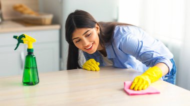 Genç bir kadın, mutfak tezgahını bir bez ve sprey temizleyici kullanarak mutlulukla temizliyor. Sarı eldivenler giyer ve parlak ve düzenli bir ortamda görevine odaklanır..