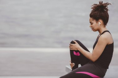 Egzersiz sırasında dizinden yaralanmış. Üzgün Afro-Amerikalı spor elbiseli, kablosuz kulaklıklı, merdivenlerde oturan ve bacağını tutan genç bir kadın.