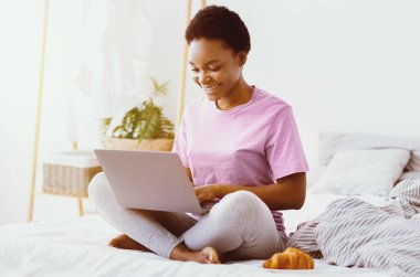 Uzaktan çalışma ve evden webinar izleme. Gülümseyen Afrikalı Amerikalı kız dizüstü bilgisayarla yatakta oturuyor, yakın tabakta kruvasan var, güneş parlıyor, boş yer var.