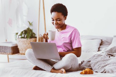 Sabah haberleri ve yazışmalar. Gülümseyen Afrikalı Amerikalı kız kahvaltısını yapar ve laptopuyla yatakta kahve içer.