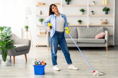 Genç bir kadın evini temizliyor, elinde yorgun bir ifadeyle paspas tutuyor. Parlak bir salonda, bitkilerin, bir kanepenin ve yakınlardaki bir kovada temizlik malzemelerinin olduğu bir salonda duruyor..