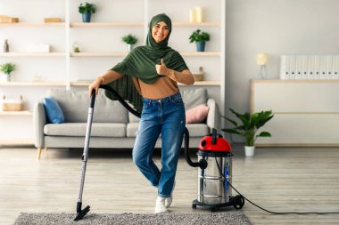 Genç bir kadın memnuniyetle oturma odasını süpürüyor. Modern kıyafetler giyiyor ve başörtüsü takıyor. Başparmağını kaldırırken halıya basıyor ve ev işlerine karşı olumlu tavrını sergiliyor..