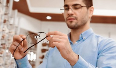 Açık mavi gömlekli bir adam optik bir dükkanda bir gözlüğü inceliyor. Tasarımlarını ve uygunluklarını dikkatle değerlendirir, çeşitli göz giysi seçenekleriyle donatılmıştır..