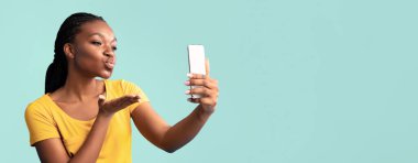 Sarı tişörtlü bir kadın fotoğraf çekerken akıllı telefonuna bir öpücük yolluyor. Arka plan canlı bir deniz mavisi, o anın neşeli atmosferine katkı yapıyor..