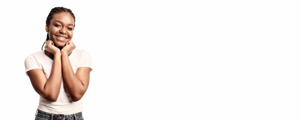 A joyful young woman stands with a beaming smile, resting her hands on her cheeks, showcasing a playful and vibrant atmosphere in a bright studio setting.