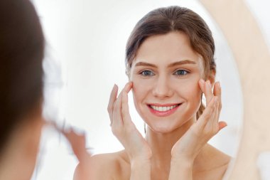 Güzel, gülümseyen bir kadın aynaya bakıyor, göz serumu uyguluyor, yaşlanmayı önleyen güzellik ürünleri konsepti