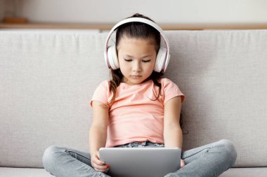 Gadget Bağımlılığı. Dijital tablet ve kablosuz kulaklık kullanan Çinli bir kız. Kanepede oturmuş çizgi film izliyor.