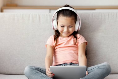 Tablet Bilgisayar kullanan Asyalı bir kız. Kablosuz kulaklık takıyor. Evde kanepede oturmuş çizgi film izliyor. Çocuk ve Gadget Konsepti