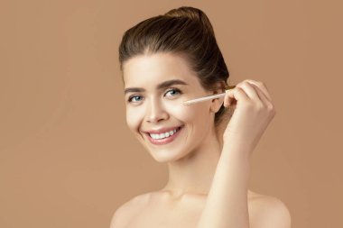 Güzellik kozmetik konsepti. Şehvetli genç bir kadın yüzüne nemlendirici serum sürüyor, kahverengi stüdyo geçmişi var. Milenyum bayanı kendini şımartıyor, cildiyle ilgileniyor.