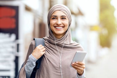 Neşeli Arap kız şehrin yanından geçiyor ve müzik dinliyor, akıllı telefon kullanıyor, boş yer var.