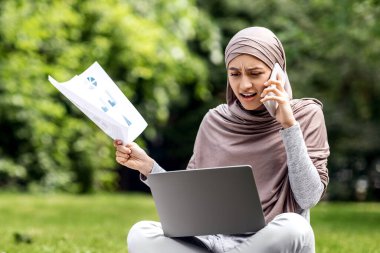 Kızgın Arap kadın parkta çalışıyor, dizüstü bilgisayar kullanıyor ve müşterilerle telefonda konuşuyor, kağıtları tutuyor, kopyalama alanı açıyor.