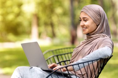 Genç tesettürlü mutlu kadın parkta bankta otururken dizüstü bilgisayar kullanıyor, açık havada çalışıyor, boş yer var.