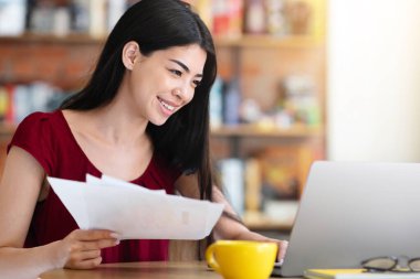 Genç Asyalı iş kadını mali raporları kontrol ediyor ve kafede dizüstü bilgisayar üzerinde çalışıyor.